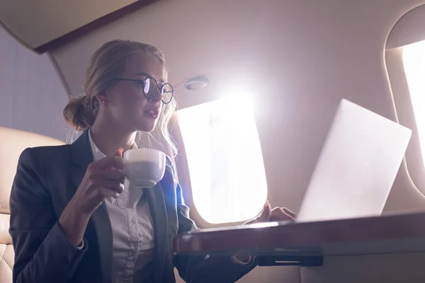 Beautiful businesswoman holding coffee cup and working on laptop in plane during business trip — Stock Photo