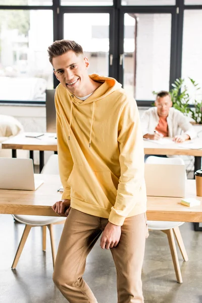 Junger lächelnder Geschäftsmann blickt in die Kamera, während er am Arbeitsplatz im Büro steht — Stockfoto