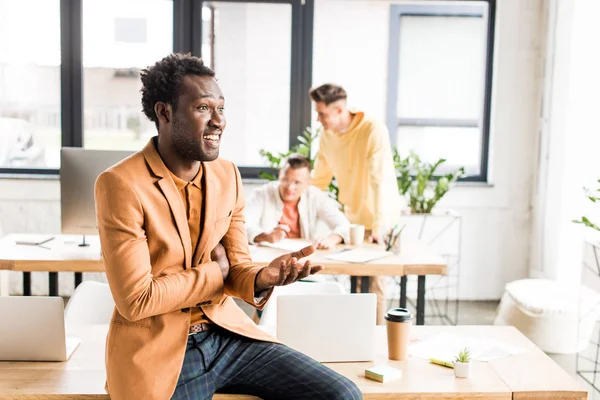 Smiling african american businessman gesturing while sitting on desk near colleagues — Stock Photo