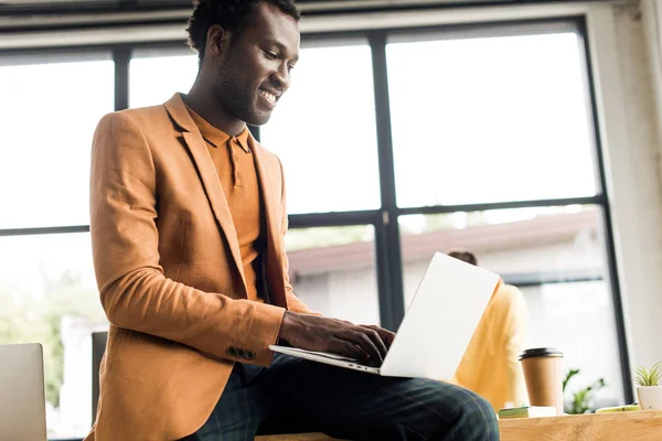 Sonriente hombre de negocios afroamericano sentado en el escritorio y utilizando el ordenador portátil - foto de stock