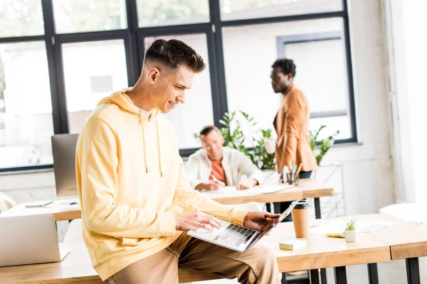 Joven hombre de negocios usando el ordenador portátil mientras está sentado en el escritorio en la oficina cerca de colegas multiculturales - foto de stock