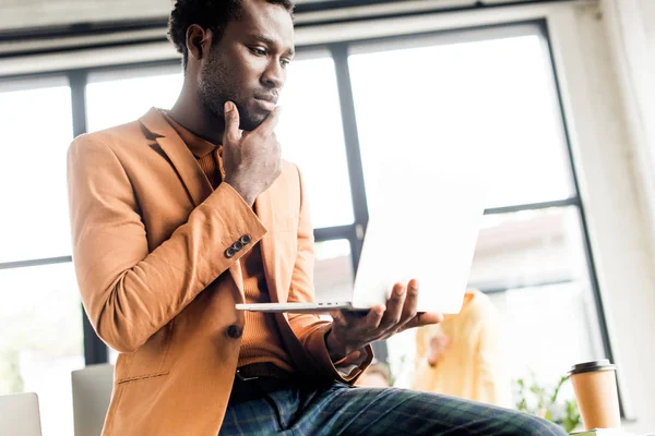 Thoughtful african american businessman sitting on desk and using laptop — Stock Photo