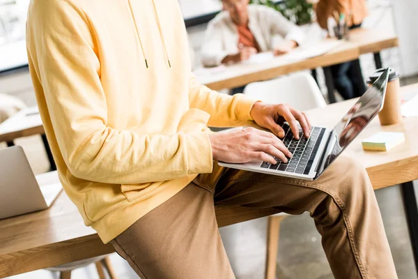 Cropped view of young businessman sitting on desk and using laptop in office — Stock Photo