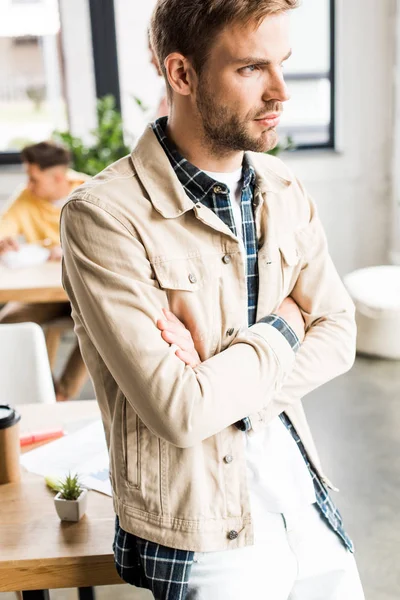 Thoughtful, young businessman looking away while standing in office with crossed arms — Stock Photo