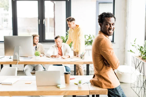 Alegre afroamericano hombre de negocios mirando la cámara cerca de colegas que trabajan en la oficina - foto de stock