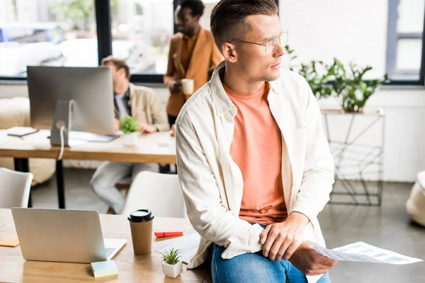 Nachdenkliche, junge Geschäftsleute, die am Schreibtisch sitzen und wegschauen, während multikulturelle Kollegen im Büro arbeiten — Stockfoto