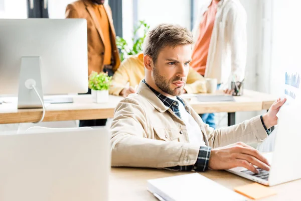 Junger, gutaussehender Geschäftsmann mit Laptop in der Nähe von Kollegen im Büro — Stockfoto