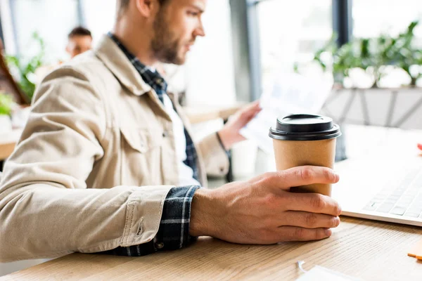 Избирательный фокус молодого бизнесмена, держащего кофе, чтобы пойти, сидя на рабочем месте в офисе — стоковое фото