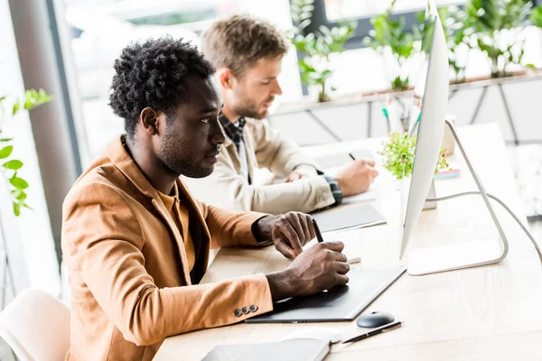 Junger afrikanisch-amerikanischer Geschäftsmann mit Tablet und Blick auf Computerbildschirm, während er neben einem Kollegen im Büro sitzt — Stockfoto