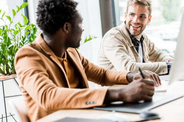 Selective focus of african american businessman talking to colleague while sitting at workplace together — Stock Photo