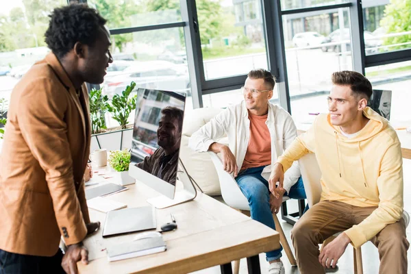 Young, multicultural businesspeople discussing startup project in office near workplace with computer monitor — Stock Photo