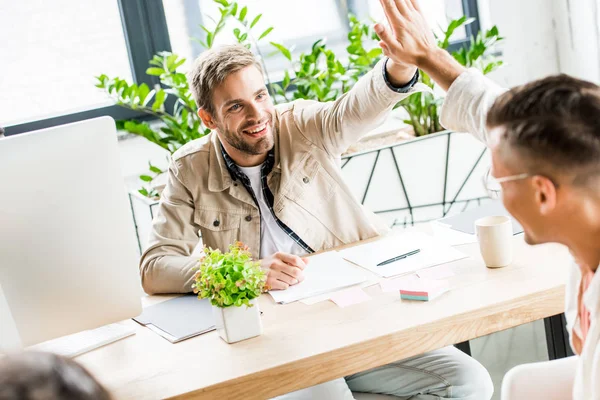 Jóvenes, hombres de negocios alegres dando cinco altos mientras está sentado en el lugar de trabajo en la oficina - foto de stock