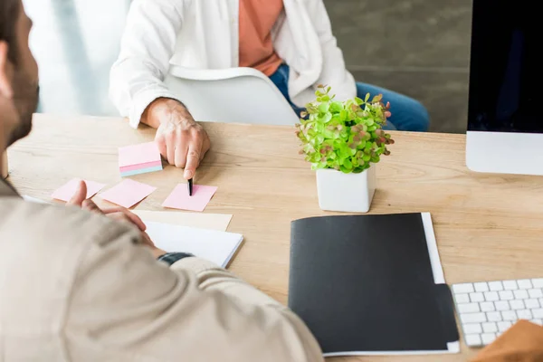 Abgeschnittene Ansicht eines Geschäftsmannes, der auf leere, klebrige Zettel zeigt, während er neben einem Kollegen am Arbeitsplatz sitzt — Stockfoto