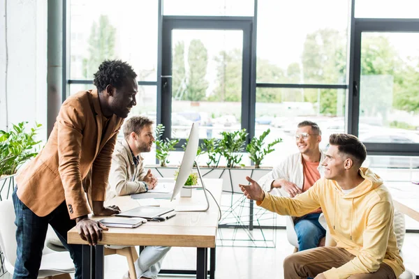 Cuatro hombres de negocios multiculturales sentados cerca de escritorio y discutiendo juntos el proyecto de startup - foto de stock