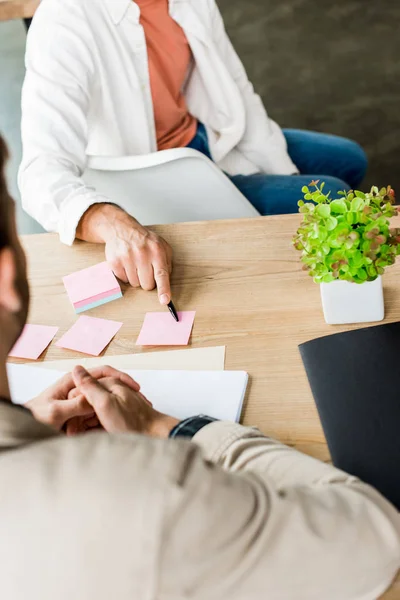 Cropped view of businessman pointing at empty sticky note while sitting near colleague at workplace — Stock Photo