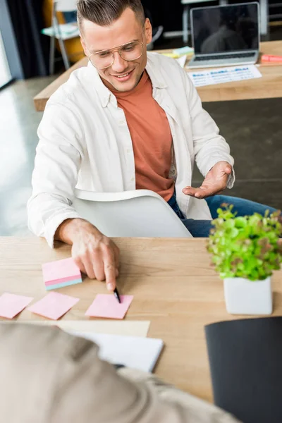 Young businessman pointing at empty sticky note while sitting near colleague at workplace — Stock Photo