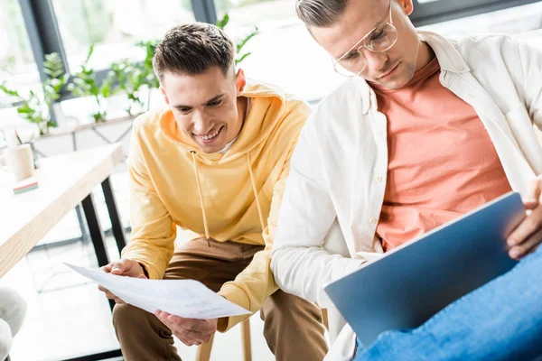 Two young businessman using laptop while sitting near colleague holding paper — Stock Photo