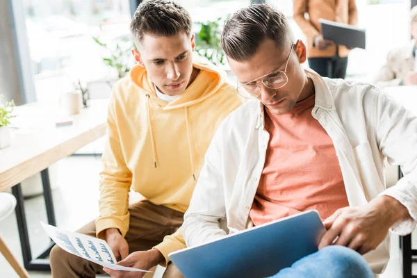 Два молодых бизнесмена, сидя рядом с коллегой, держат бумагу с графиками и диаграммами. — стоковое фото