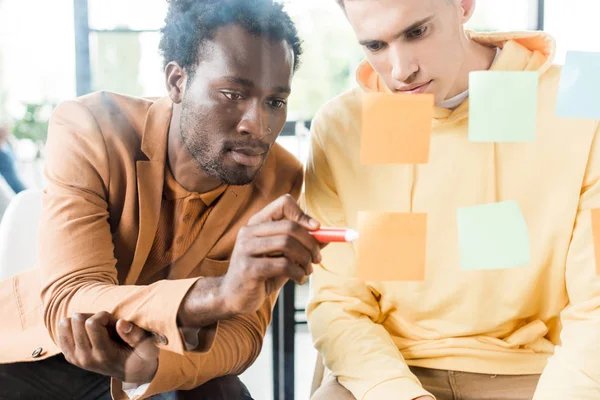 Joven hombre de negocios afroamericano escribir en nota adhesiva fija en tablero de vidrio mientras está sentado cerca de colega - foto de stock