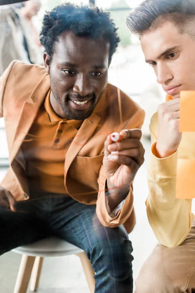 Young african american businessman writing on glass board while sitting near colleague — Stock Photo