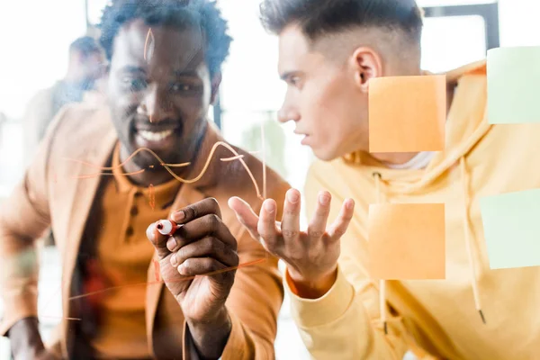 Smiling african american businessman writing on glass board while sitting near colleague — Stock Photo