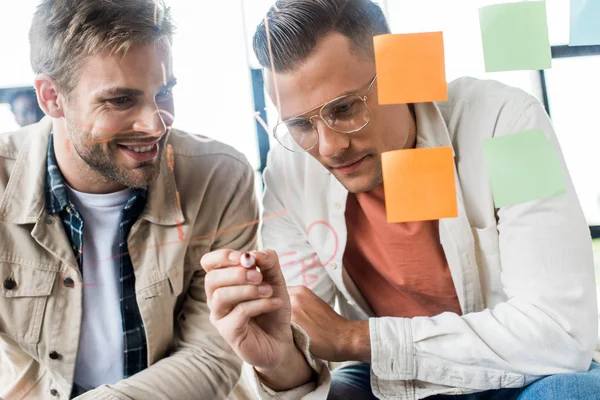 Young businessman writing on glass board while working with colleague in office — Stock Photo