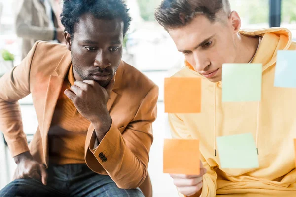 Deux jeunes hommes d'affaires multiculturels regardant des notes collantes fixées sur un panneau de verre au bureau — Photo de stock