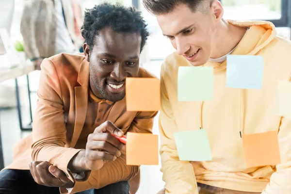 Sonriente hombre de negocios afroamericano escribiendo en nota adhesiva mientras está sentado cerca de un colega en la oficina - foto de stock