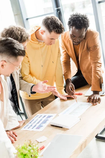 Four multicultural businesspeople analyzing papers with graphs and charts while working on startup project together in office — Stock Photo