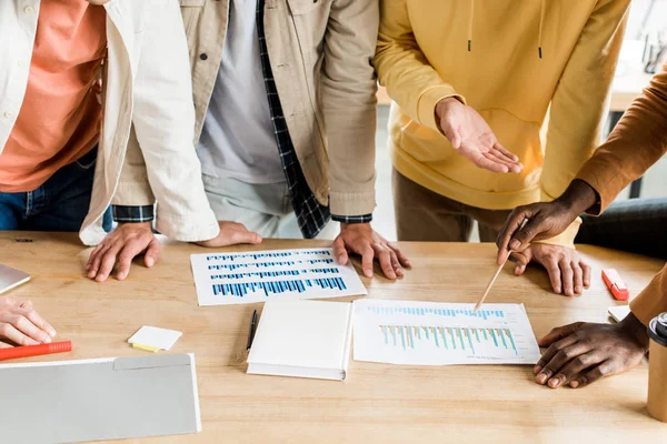 Vista recortada de cuatro empresarios multiculturales que analizan documentos con gráficos y gráficos mientras trabajan juntos en el proyecto de startup en la oficina - foto de stock