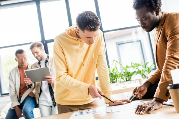 Jóvenes empresarios multiculturales analizando documentos con gráficos y gráficos mientras trabajan juntos en el proyecto de startup en la oficina - foto de stock
