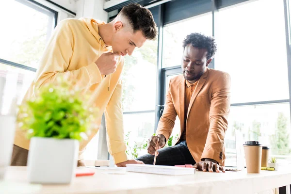 Afrikanisch-amerikanischer Geschäftsmann sitzt auf Schreibtisch neben Kollege, der in der Nähe im Büro steht — Stockfoto