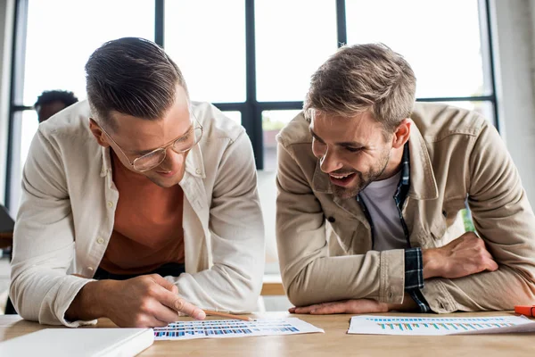 Двоє молодих усміхнених бізнесменів аналізують документи з графіками та діаграмами під час роботи над стартап-проектом разом в офісі — стокове фото