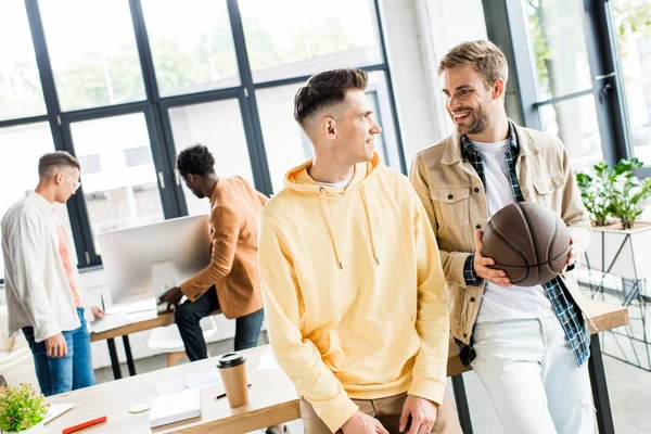 Smiling businessman holding volleyball while talking to colleague in office — Stock Photo