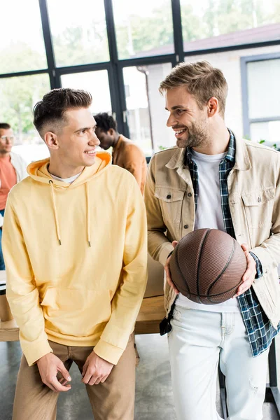 Sonriente hombre de negocios sosteniendo voleibol mientras habla con su colega en la oficina - foto de stock