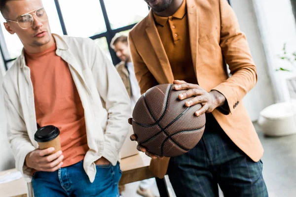 Cropped view of african american businessman holding volleyball while standing near colleague with coffee to go in office — Stock Photo
