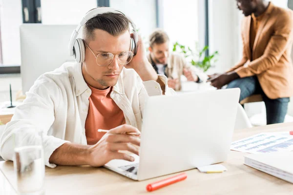 Joven hombre de negocios en auriculares usando el ordenador portátil mientras trabaja cerca de colegas multiculturales - foto de stock