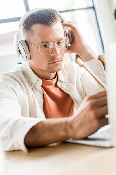 Joven, hombre de negocios reflexivo sentado en los auriculares y el uso de la computadora portátil en la oficina - foto de stock