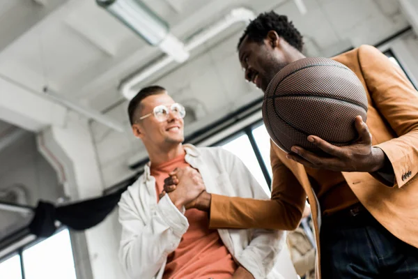 Low angle view of african american businessman holding volleyball and shaking hands with colleague in office — Stock Photo
