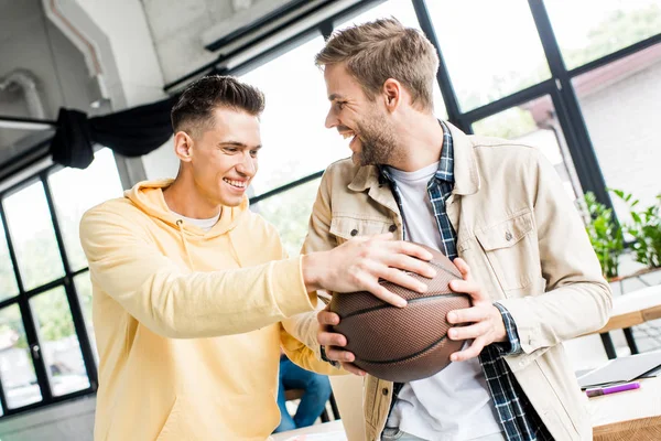 Young cheerful businessman taking volleyball from hands of smiling colleague in office — Stock Photo