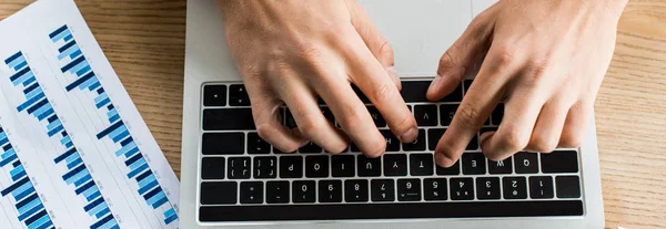 Обрезанный вид бизнесмена, работающего на ноутбуке рядом с бумагой с графиками и графиками, панорамный снимок — стоковое фото