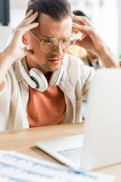 Jeune homme d'affaires coûteux regardant ordinateur portable tout en étant assis avec des écouteurs sur le cou — Photo de stock