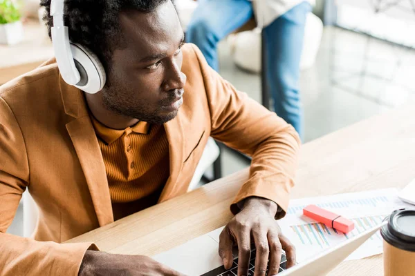 Hombre de negocios afroamericano reflexivo en los auriculares usando el ordenador portátil en la oficina - foto de stock