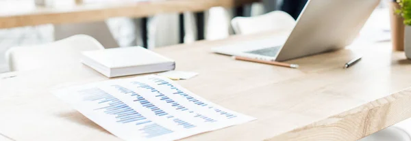 Panoramic shot of desk with laptop, notebook and paper with graphs and charts in office — Stock Photo