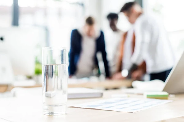 Selective focus of glass with water on desk near businesspeople standing at workplace in office — Stock Photo