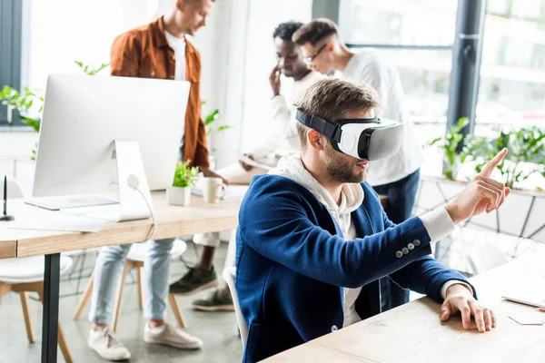 Молодой бизнесмен, использующий наушники vr и трогающий что-то пальцем в то время как мультикультурные коллеги работают в офисе — стоковое фото