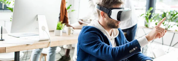 Panoramic shot of young businessman touching something with finger while using vr headset in office — Stock Photo