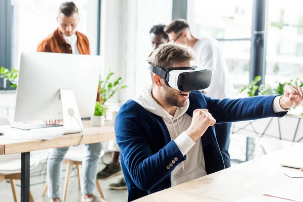 Joven empresario imitando lucha mientras usa auriculares vr, y colegas multiculturales que trabajan en la oficina - foto de stock