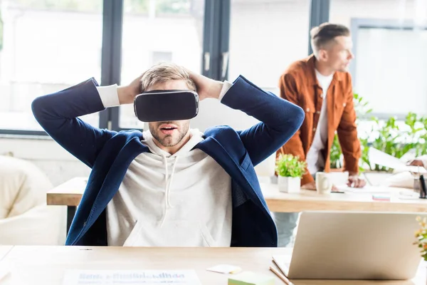 Giovane uomo d'affari utilizzando auricolare vr e tenendosi per mano dietro la testa mentre seduto sul posto di lavoro in ufficio — Foto stock