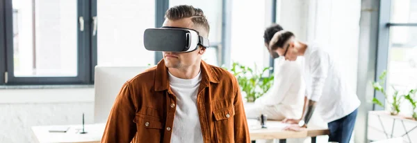 Panoramic shot of young businessman using vr headset while multicultural colleagues working in office — Stock Photo